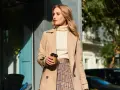 Elegancka kobieta w beżowym płaszczu, kremowym golfie i spódnicy w kratę, z kubkiem kawy w dłoni. Inspiracja na stylizacje do beżowego płaszcza.
