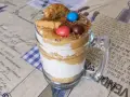 Warstwowy deser banoffee w szklance, ozdobiony ciasteczkiem, cukierkami i wiórkami czekolady.