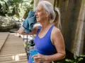 Femme d'âge mûr s'essuyant le visage avec une serviette bleue, tenant une bouteille d'eau. Elle fait du sport malgré la canicule.