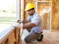 Un ouvrier en casque jaune travaille sur le remplacement de fenêtres dans une maison en construction, mesurant le cadre.