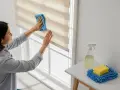 Mujer limpiando estores enrollables con un paño azul. A su lado, un spray y una esponja.