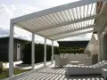 Un moderno porche de obra con techo de lamas ajustables, mobiliario de exterior blanco y un camino de madera que conduce a una cabaña.