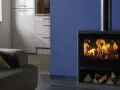 Estufa de leña moderna con fuego encendido y leña apilada, junto a un sofá y muebles de cocina.