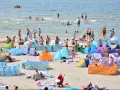Wczasy pod gruszą na zatłoczonej plaży. Ludzie kąpią się w morzu, opalają na piasku i odpoczywają pod kolorowymi parawanami.
