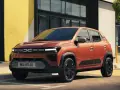 Pomarańczowy Dacia Spring stoi przed budynkiem z żółtą ścianą.