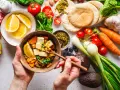 Zdrowa dieta na stawy: miska pełna soczewicy, tofu, warzyw i cytryny. Obok składniki na pyszne danie: pomidory, marchew, awokado, czosnek, cebula, jagody, pesto i chlebki pita.
