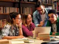 Vier Studierende lernen in der Bibliothek. Ein Mann, der wie ein seelsorger quereinsteiger wirkt, erklärt etwas am Laptop.