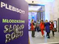 Plakat "PLEBISCYT Młodzieżowe Słowo Roku" z lwem w koronie. W tle młodzież wchodzi do budynku.