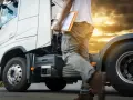 Kierowca z teczką zbliża się do białej ciężarówki. Zachód słońca i chmury tworzą dramatyczne tło dla tej sceny, która może być początkiem ważnej podróży lub zakończeniem dnia pracy na konwencja tir.