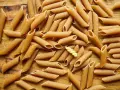 Mnóstwo makaronu pełnoziarnistego penne na drewnianym blacie. Idealny wybór dla dbających o linię, bo makaron pełnoziarnisty kcal ma mniej!