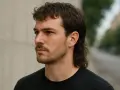 Mężczyzna z charakterystyczną fryzurą mullet, z krótkimi włosami z przodu i dłuższymi z tyłu, patrzy w bok.