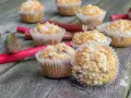 Pyszne muffinki z rabarbarem, posypane kruszonką, leżą na drewnianym blacie obok świeżych łodyg rabarbaru.