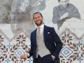 Elegancki mężczyzna w granatowym garniturze i kamizelce, idealny przykład, jak się ubrać na ślub cywilny. Uśmiecha się na tle malowidła.