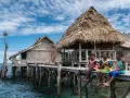 Trzech chłopców siedzi na drewnianym pomoście przy tradycyjnych chatach na wodzie w Raja Ampat.