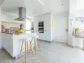 Moderna cocina blanca y gris claro con isla central, taburetes y frutas frescas.