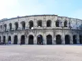 The ancient Roman amphitheater in Nîmes, France, stands majestically under a clear blue sky. Tourists gather in the plaza, ready to explore this historic marvel.