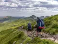 Randonnée sur le Puy Mary, des marcheurs explorent un sentier rocailleux bordé de verdure, avec des montagnes ondulantes en arrière-plan sous un ciel nuageux.