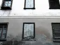 Stara elewacja z widocznymi pęknięciami i zaciekami. Okna z żaluzjami i jedno wybite.
