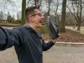 Mężczyzna w okularach trzyma but Merrell przed otwartymi ustami w parku.