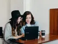 Dwie młode kobiety pracują na laptopie, jedna w kapeluszu, druga w dżinsowej koszuli. Ich wspólny projekt to passe recent.