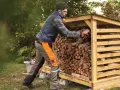 Mężczyzna w ogrodowych ubraniach układa drewno opałowe w drewnianej wiacie.