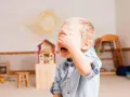 Płacz 3-latka, zasłaniającego twarz ręką, może wskazywać na niepokojące zachowania. W tle pokój zabaw z domkiem dla lalek i rysunkiem słońca.