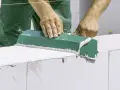 Ręce budowlańca w zielonym fartuchu rozprowadzają zaprawę przy murowaniu ścian z białych bloczków.