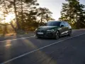 Un'auto verde scuro, un segmento premium, sfreccia su una strada panoramica al tramonto, con alberi sfocati sullo sfondo.