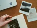 Dłoń układa zdjęcie na albumie, obok inne fotografie i laptop. Idealne miejsce, gdzie przechowywać zdjęcia z telefonu.