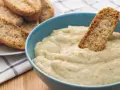 Un trozo de pan crujiente se sumerge en un cremoso baba ganoush en un cuenco azul. A un lado, hay una pila de pan tostado.