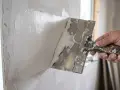 En hand bredspacklar en vägg med vit spackel. Verktyget är en bredspackel med trähandtag.