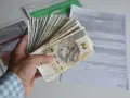 Dłoń trzyma plik banknotów 200 zł. W tle dokumenty, być może dotyczące tego, ile godzin to 0.5 etatu.