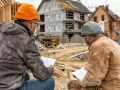 Deux hommes en casque discutent des plans sur un chantier. L'un d'eux, un artisan qui ne vient pas faire les travaux, semble expliquer quelque chose.