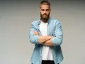 Uomo con barba e capelli biondi, camicia azzurra aperta su t-shirt bianca, braccia conserte. Look trasandato ma curato.