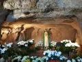 A statue of Mary stands in a grotto, surrounded by lush white flowers. The cave walls are illuminated, creating a serene atmosphere, reminiscent of the three fountains of grace.