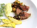 Rehleber zubereiten: zarte Scheiben Rehleber mit gebratenen Zwiebeln, Kartoffelpüree, gebratenen Kartoffelspalten und frischen Kräutern auf einem weißen Teller.
