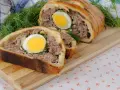 Une tranche de pâté de Pâques, garnie d'un œuf dur, est présentée sur une planche en bois, avec de l'aneth frais.
