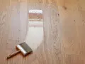 Breite Pinsel mit Holzgriff trägt Lack auf Parkett auf. Der Prozess, das Parkett zu versiegeln, zeigt den Unterschied zwischen unbehandeltem und frisch lackiertem Holz.