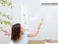 Mujer ajusta el fan speed del aire acondicionado con el control remoto, rodeada de una planta con flores rosas.