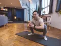 Homme en tenue de sport effectuant un étirement pubalgie sur un tapis de yoga dans un salon moderne.