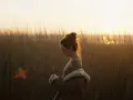 Une jeune femme, le regard baissé, se tient dans les hautes herbes au crépuscule. La lumière dorée du soleil couchant illumine ses cheveux et le paysage.