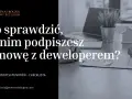 Dłoń podpisująca umowę deweloperską, klucze, mały domek i laptop.