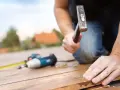 Budowa tarasu z drewna. Mężczyzna wkręca śrubę młotkiem, obok leży wkrętarka i miarka.
