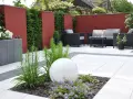 Moderner Garten ohne Rasen mit roten Mauern, weißen Terrassenplatten, dunklen Lounge-Möbeln und einer leuchtenden Kugel.