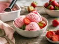 Schneebälle aus schnellem Erdbeereis, garniert mit frischen Erdbeeren und Minze, auf einem Holztisch.