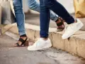 Dwie osoby w jeansach i modnych butach, z torbami na zakupy w rękach, idą chodnikiem. Jedna ma czarne sandały na obcasie, druga białe sneakersy.