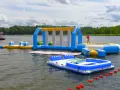 Kolorowe dmuchane atrakcje dla dzieci na jeziorze: zjeżdżalnia, trampolina i dmuchane wyspy. W tle las i łódka.