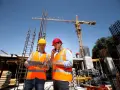 Dwóch budowlańców w kamizelkach odblaskowych omawia projekt na placu budowy. Jeden z nich trzyma tablet, drugi rozmawia przez telefon. W tle widać dźwig i zbrojenia, co sugeruje, że posiadają odpowiednie uprawnienia UDT.