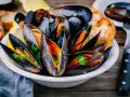 Mejillones en salsa, servidos en un cuenco blanco con perejil fresco, rodajas de limón y pan tostado.