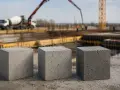 Trzy betonowe kostki, symbolizujące beton C30/37 proporcje, na tle budowy z betoniarką i dźwigiem.